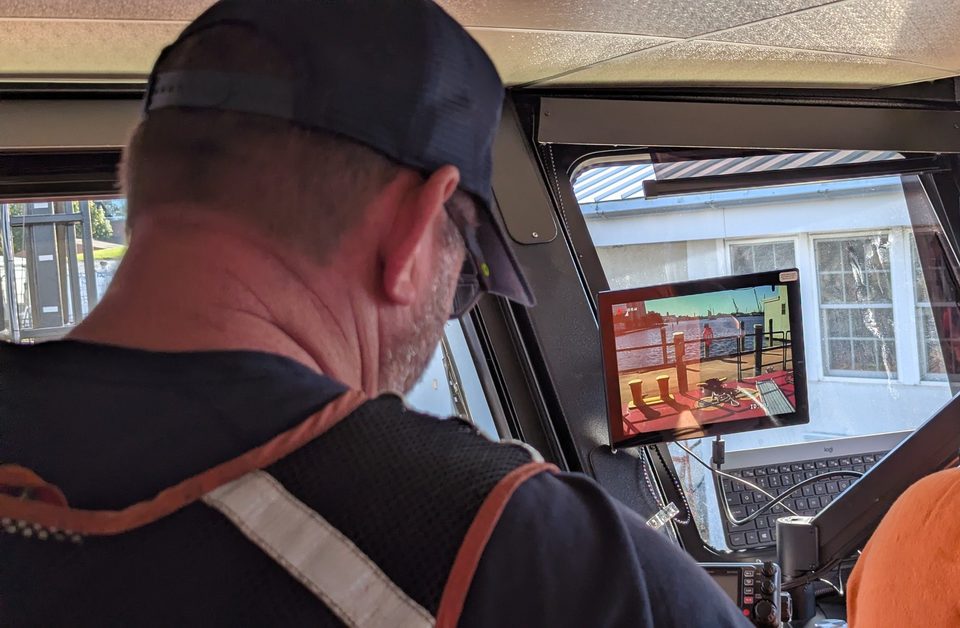 Operator in a vessel wheelhouse watching the HoistCam monitor, which shows the barge deck below the crane