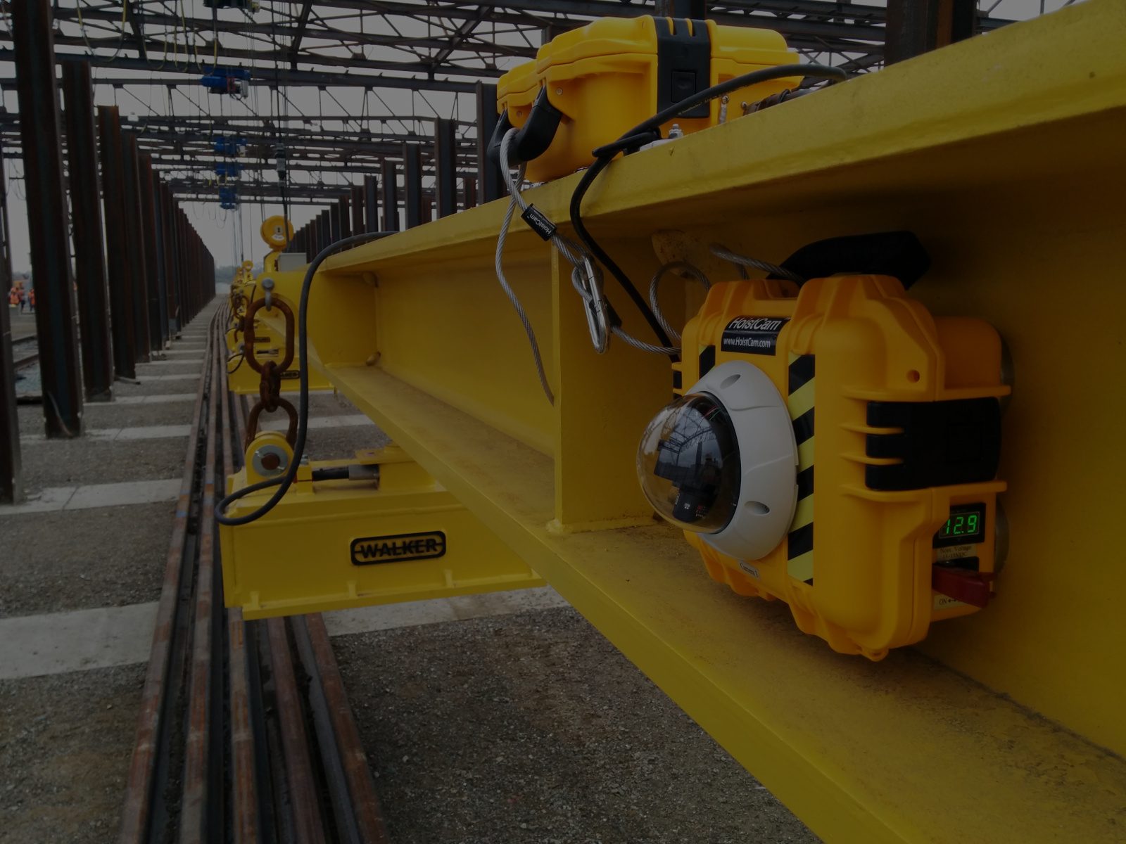 A HoistCam camera in a yellow case, mounted on the beam of an overhead crane in a steel yard